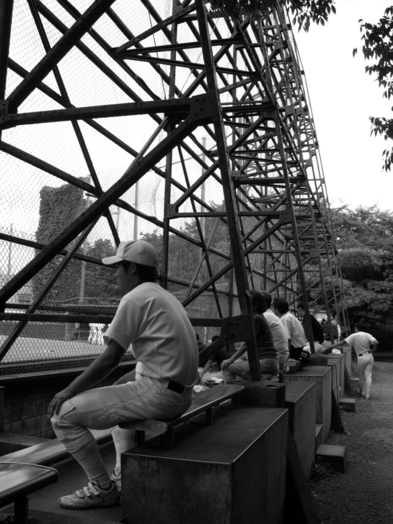 Spectateurs et joueurs de baseball derrière une grille observant un match ou un entraînement au Japon.