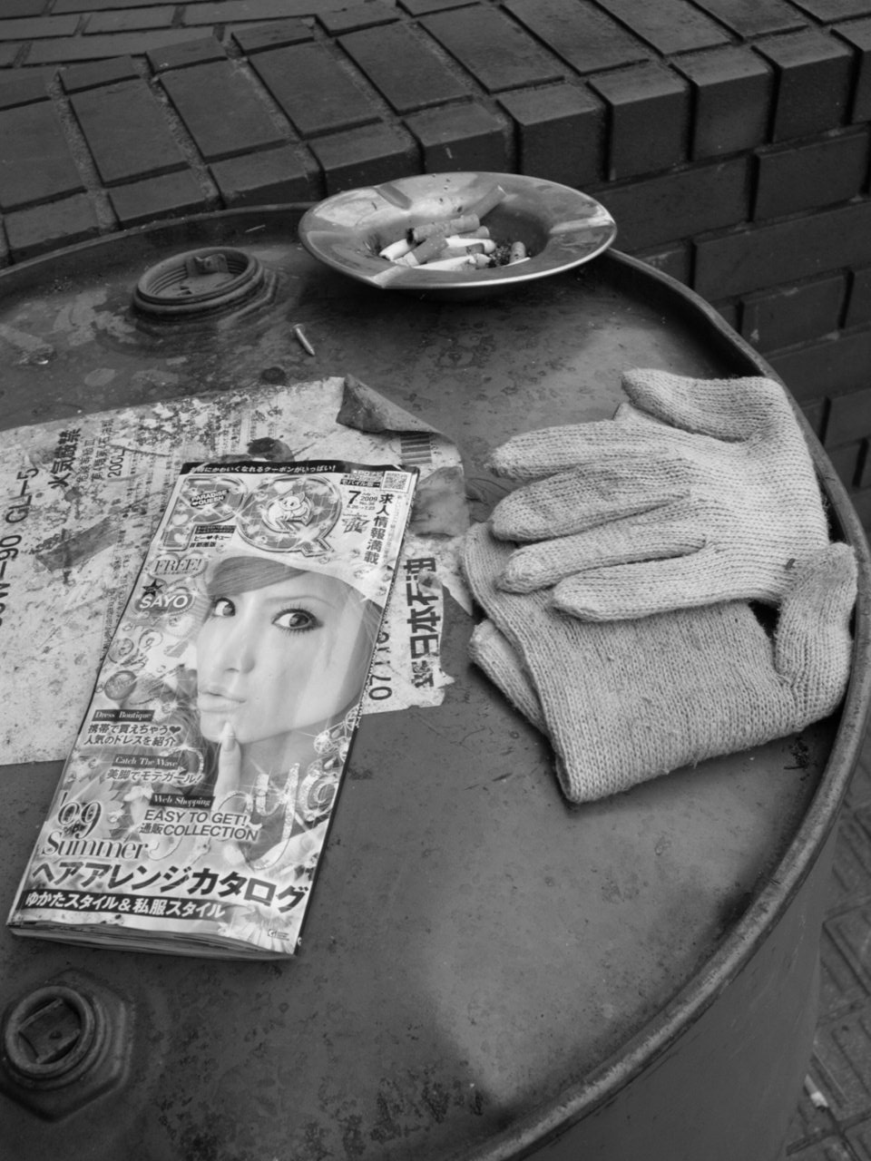 Nature morte sur un bidon métallique : cendrier, catalogue japonais et gants de travail.