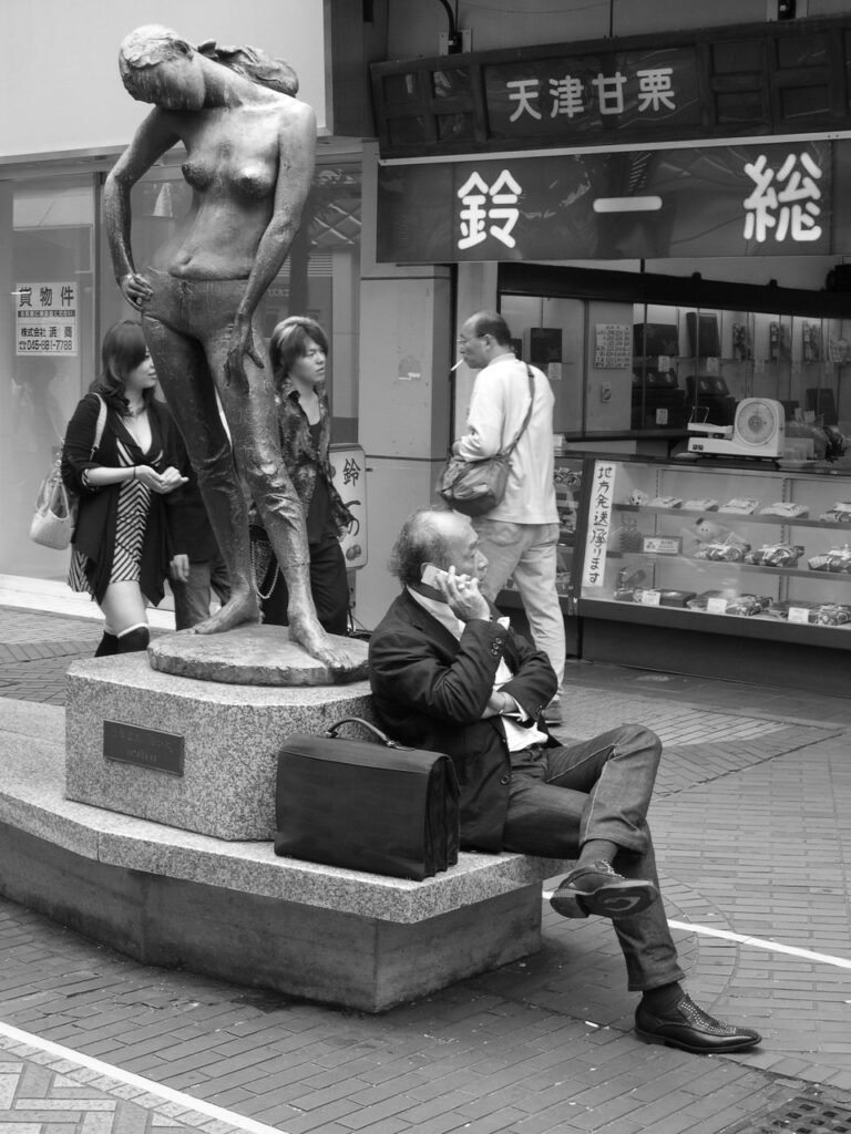 Un homme téléphone au pied d'une statue représentant une jeune femme aux seins nus, dans une rue japonaise.