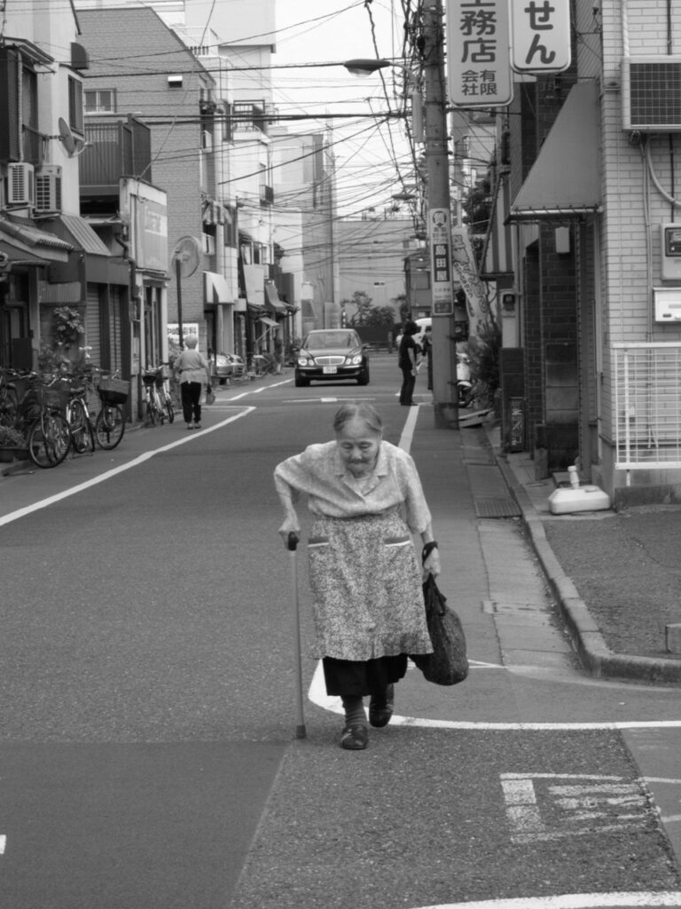 Femme âgée marchant avec une canne et un sac de courses dans une rue au Japon.
