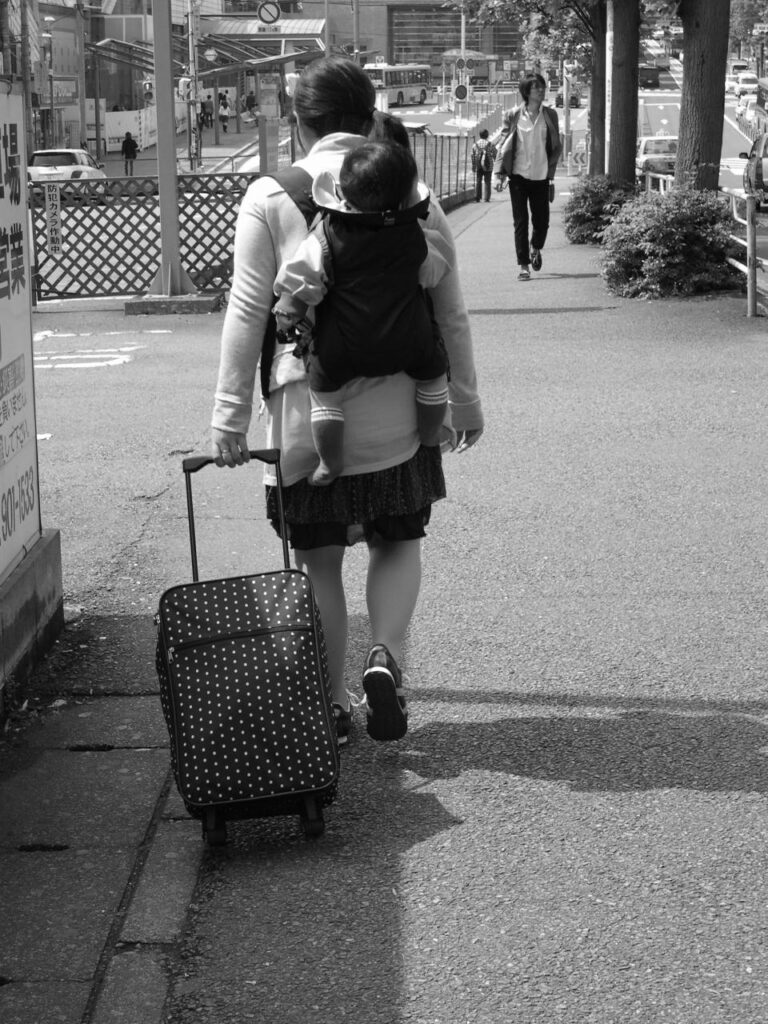 Jeune femme portant sa petite fille sur le dos et tirant une valise, tandis qu'un jeune homme s'avance en face d'elle.