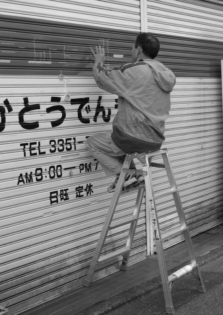 Un homme assis sur un escabeau inscrit des informations sur le rideau métallique d'un magasin au Japon.