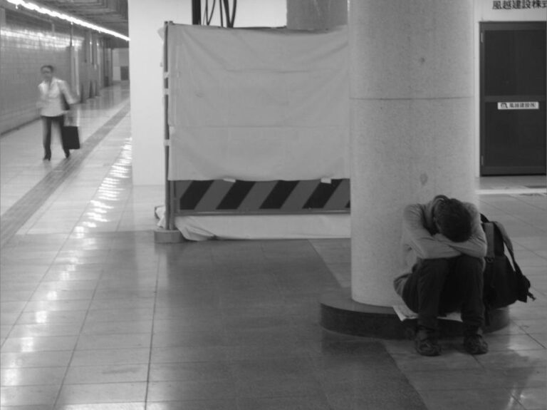 Homme assis dans un couloir de métro, la tête entre les bras, tandis qu’une femme approche avec une valise.