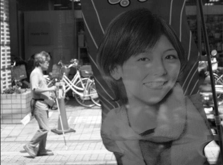 Scène de rue au Japon avec une bannière publicitaire représentant une jeune femme souriante et un passant muni d’une canne blanche.