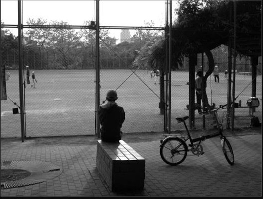 Au Japon un enfant attend, immobile, au bord d'un terrain de baseball. A côté de lui son vélo repose sur sa béquille.