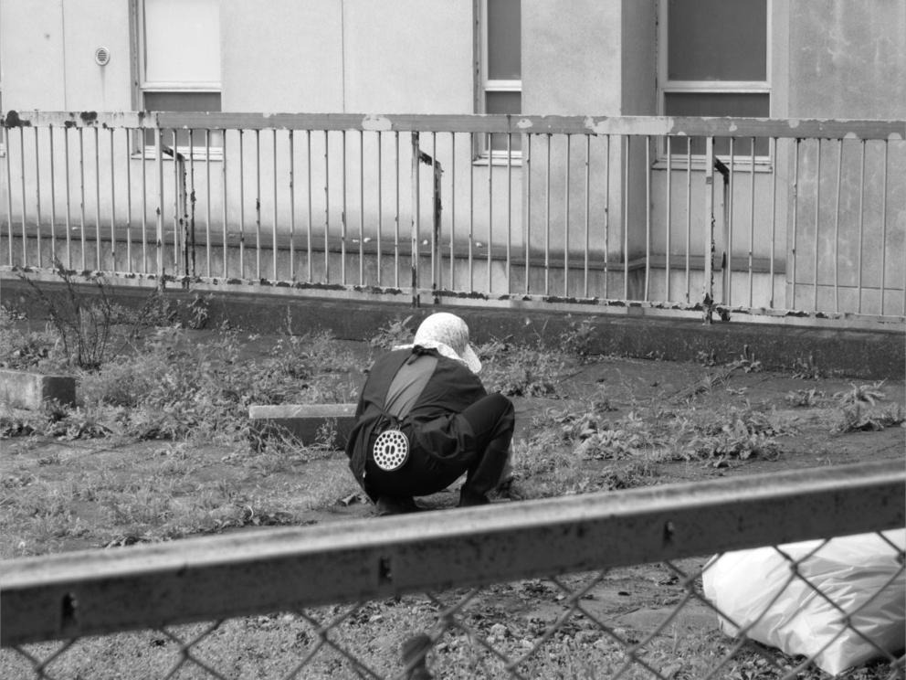 Au Japon, une femme accroupie de dos travaille la terre dans un jardin en friche devant un immeuble.