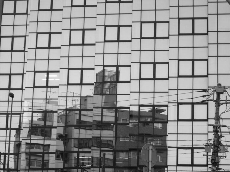 Reflet déformé d'une résidence vieillissante dans la façade vitrée d'un immeuble neuf, formant des fragments répétés et décalés.