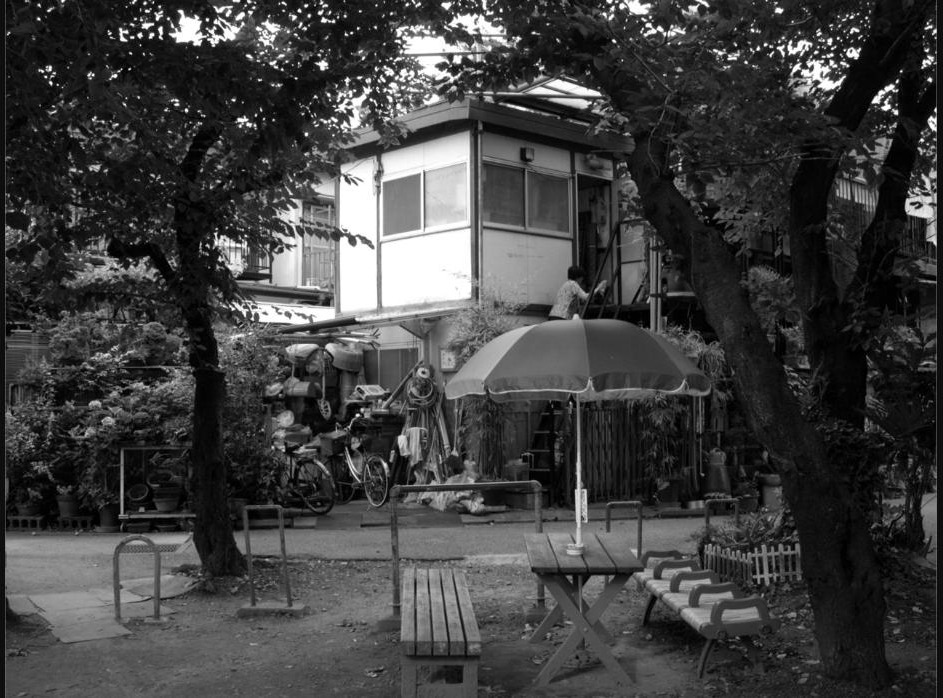 Quartier résidentiel japonais avec une maison entourée de végétation et une femme montant un escalier extérieur.