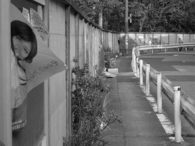 Passage étroit entre une vieille rambarde et une palissade en béton au Japon, avec une affiche publicitaire partiellement décollée.