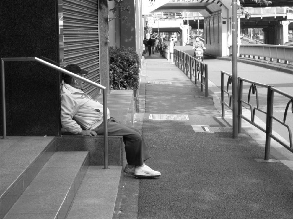 Homme âgé adossé au rideau métallique d'un commerce, observant les passants au Japon.