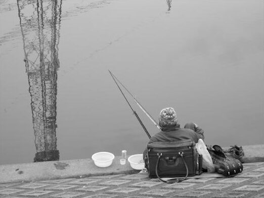 Pêcheur vu de dos, sa canne plonge dans la rivière. A gauche, le reflet dans l'eau d'une tour électrique métallique.