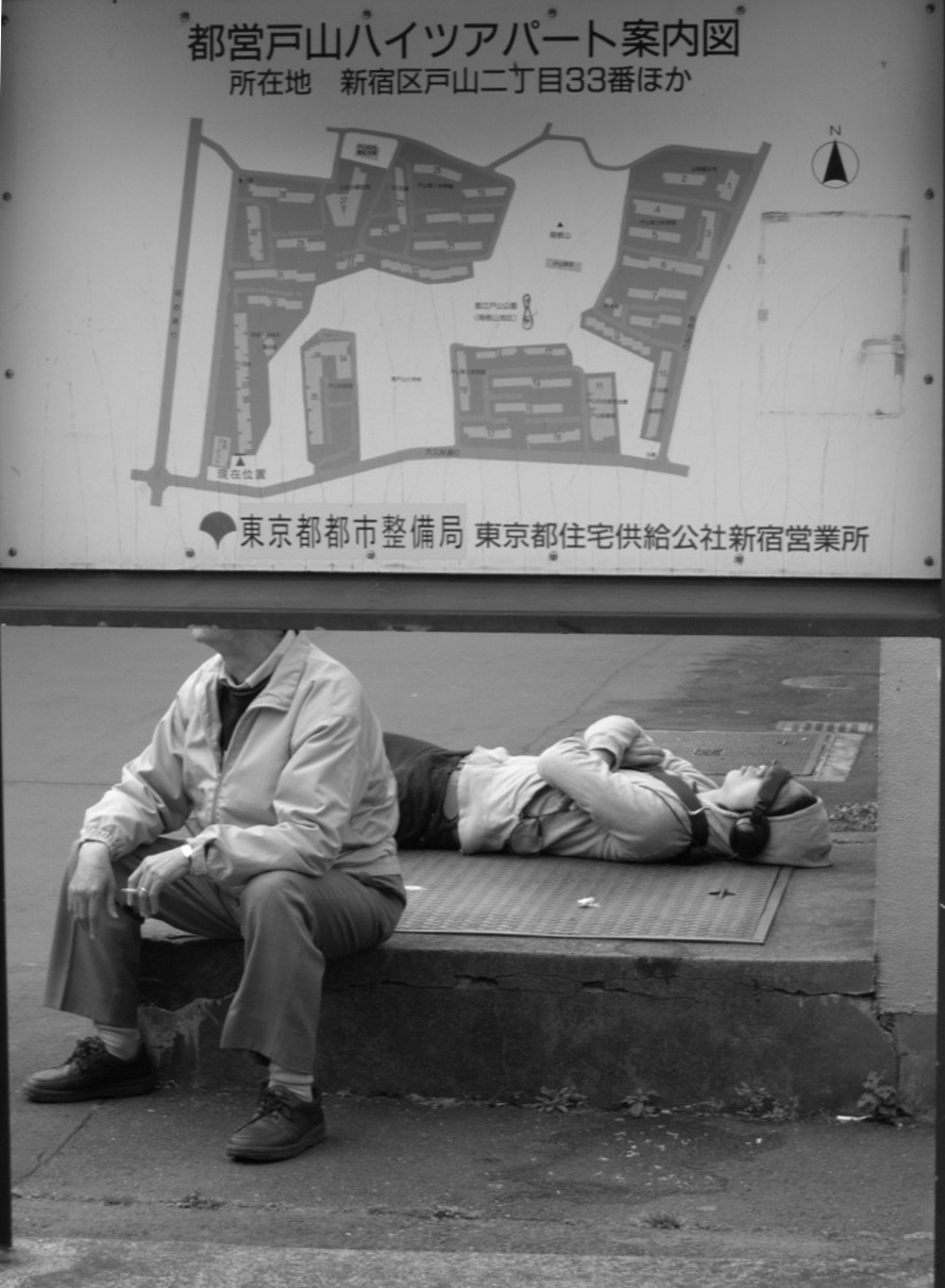 Un homme et un jeune homme font une pause sous le plan d'une résidence. L'un fume une cigarette tandis que l'autre, allongé, porte un casque sur les oreilles.