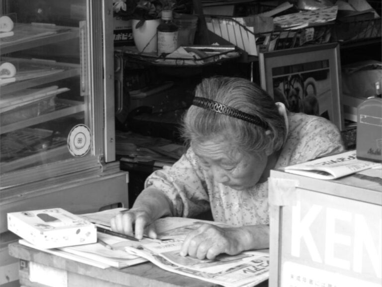 Femme âgée tenant une petite boutique au Japon, penchée sur un journal ou un carnet.