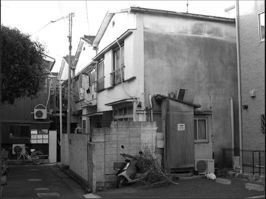 Arrière-cour japonaise en noir et blanc, bordée de petites maisons urbaines, avec vélos, scooter et détails du quotidien.